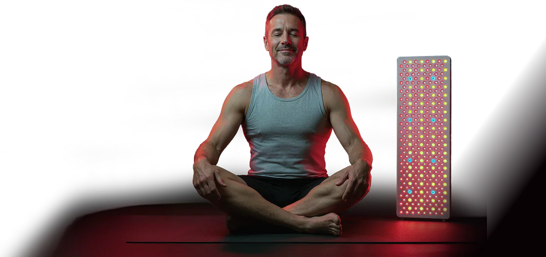 A man in a gray tank top sits cross-legged on a yoga mat, eyes closed, beside a glowing red light therapy panel. The setting is calm and meditative.