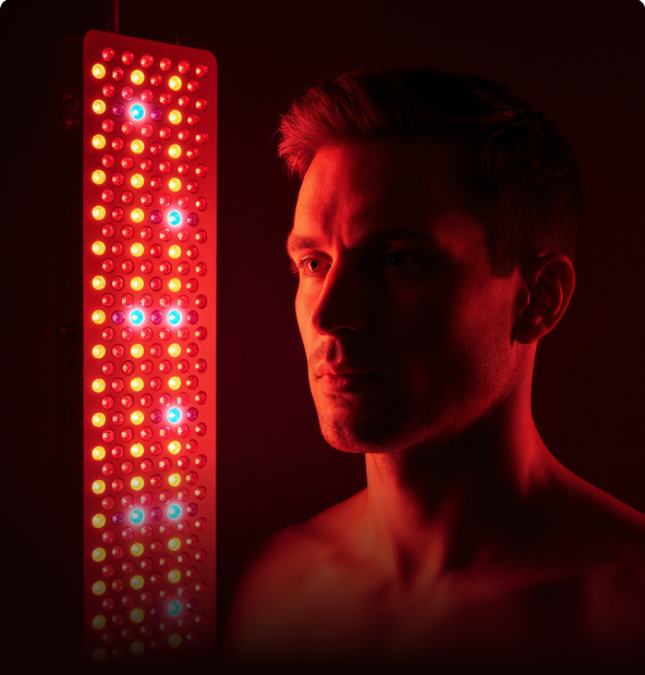 Man stands next to a glowing red LED therapy light panel in a dark setting. The red and blue lights cast an intense, focused expression on his face.